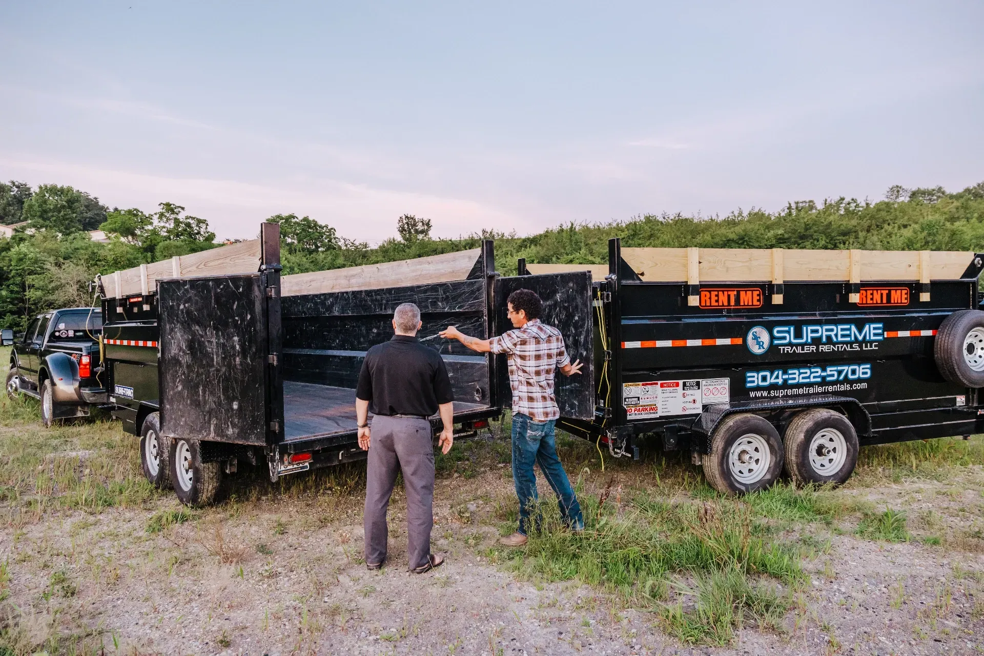 Two men inspect open dump trailers, possibly for a landscaping or construction project. Trailers are black with company branding.