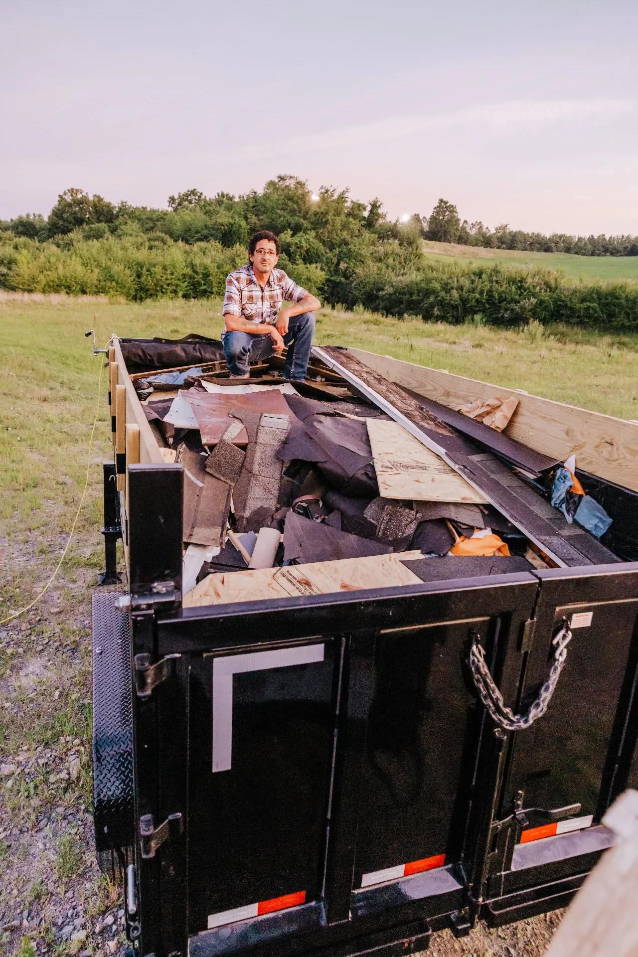 Man sits on a filled trailer with debris in a rural setting. He wears a plaid shirt, jeans, and boots; field in the background.
