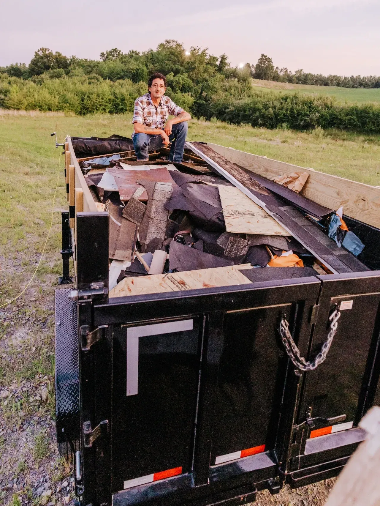 Man sits on a loaded trailer full of debris in a rural setting with a green field and trees in the background.