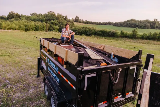 Person sits atop a loaded black trailer with wood planks, in a grassy field under a dusky sky, with a tree line in the background.
