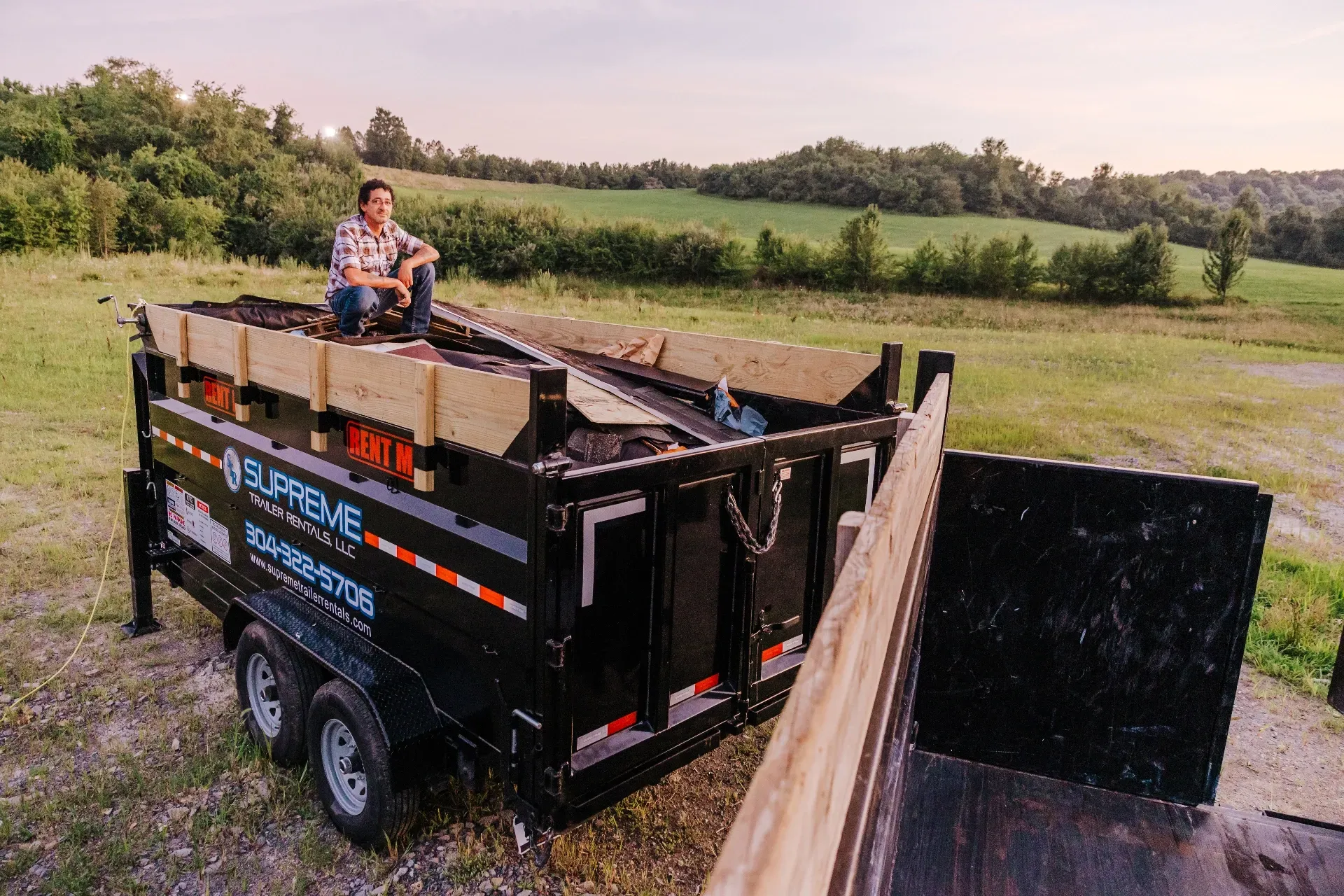 A person sits on the edge of a black Supreme dump trailer in a field. They wear a plaid shirt, and the trailer bed is filled with material.