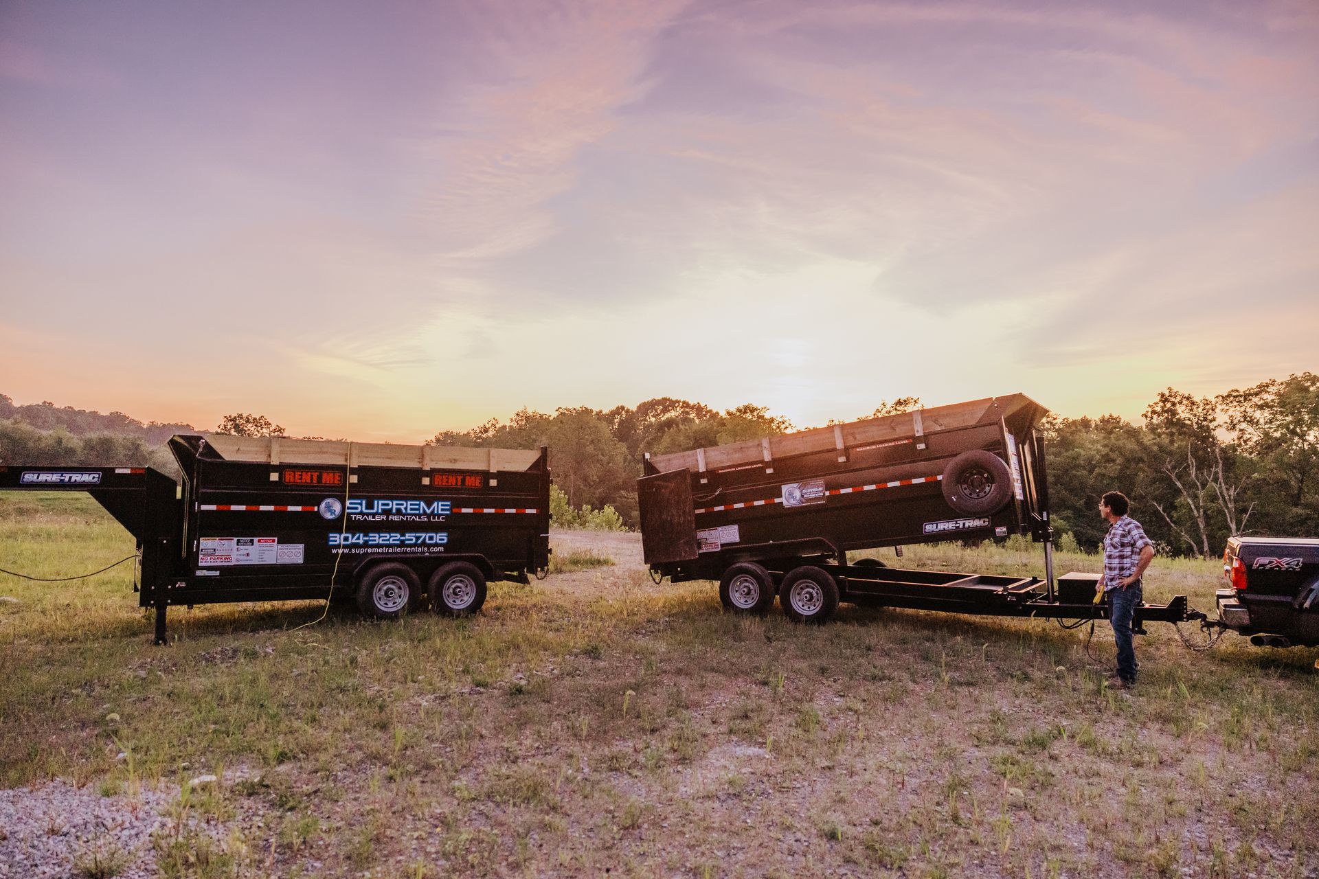 Two black dump trailers in a field, one tilted up. A man stands near the second trailer, with trees and a sunset in the background.