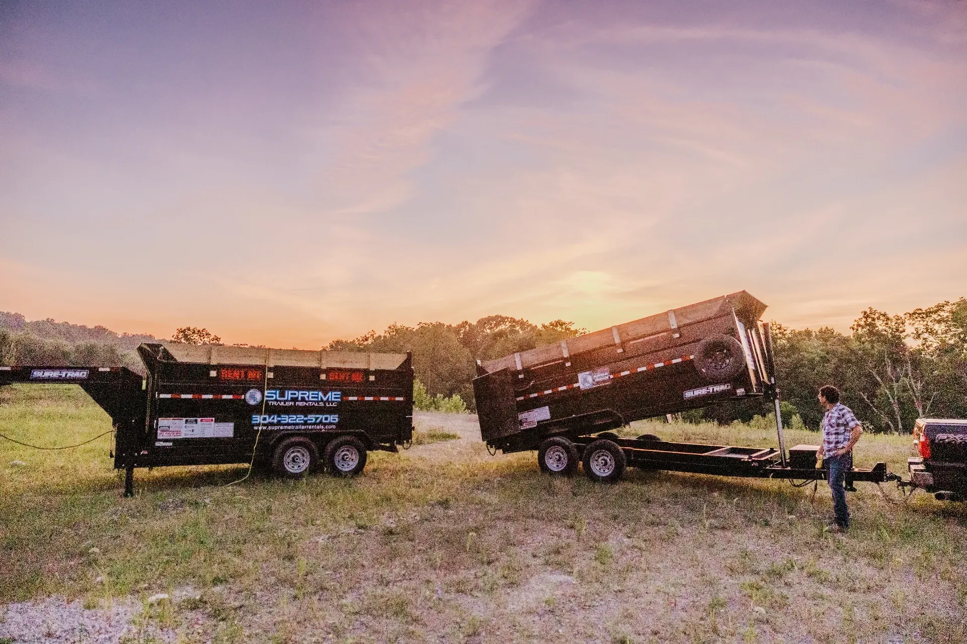 Two black dump trailers on grass field; one raised. Man in plaid shirt stands near. Sunset in the background.