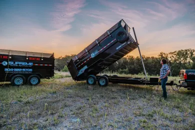 Man standing next to a black dump trailer, tilted upward, with a second trailer nearby at sunset.