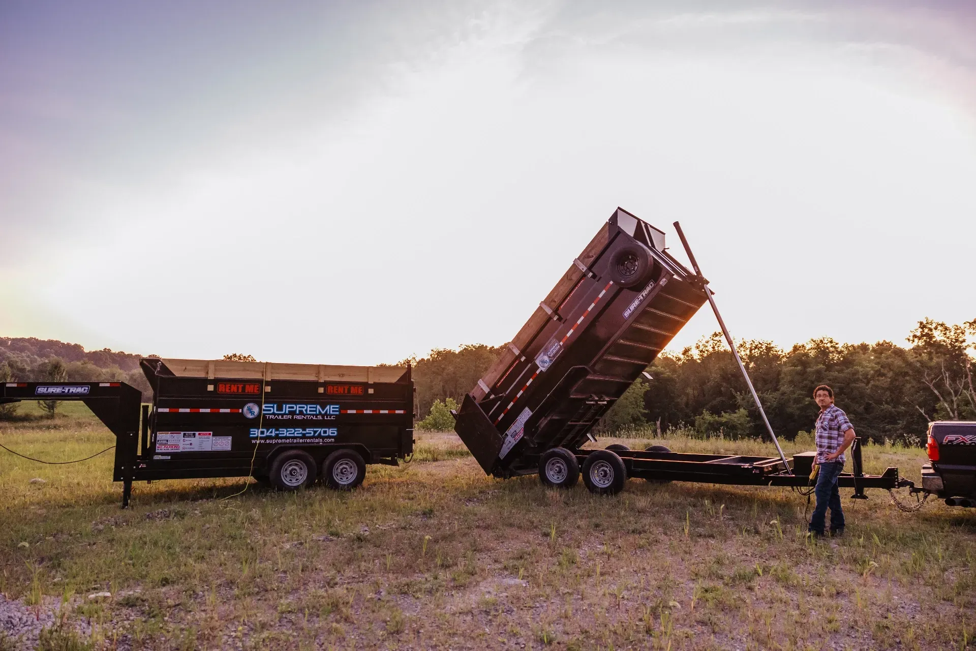 A man stands next to a black dump trailer in a grassy field as the bed is raised. A setting sun provides an orange glow.