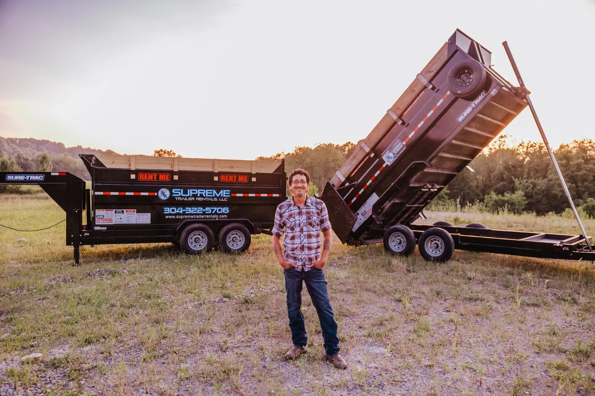Man standing in front of a black dump trailer with its bed raised, in a field with trees in the background.