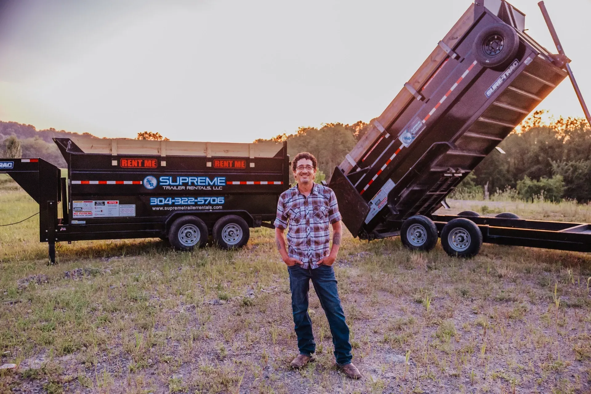Man stands in front of a large black dump trailer, one side lifted, on a grassy field with trees in the background.