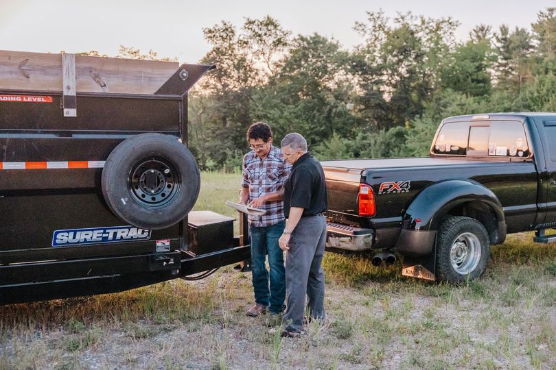 Two men standing near a black truck with a trailer. One man is looking at a phone, while the other looks on, in a field.