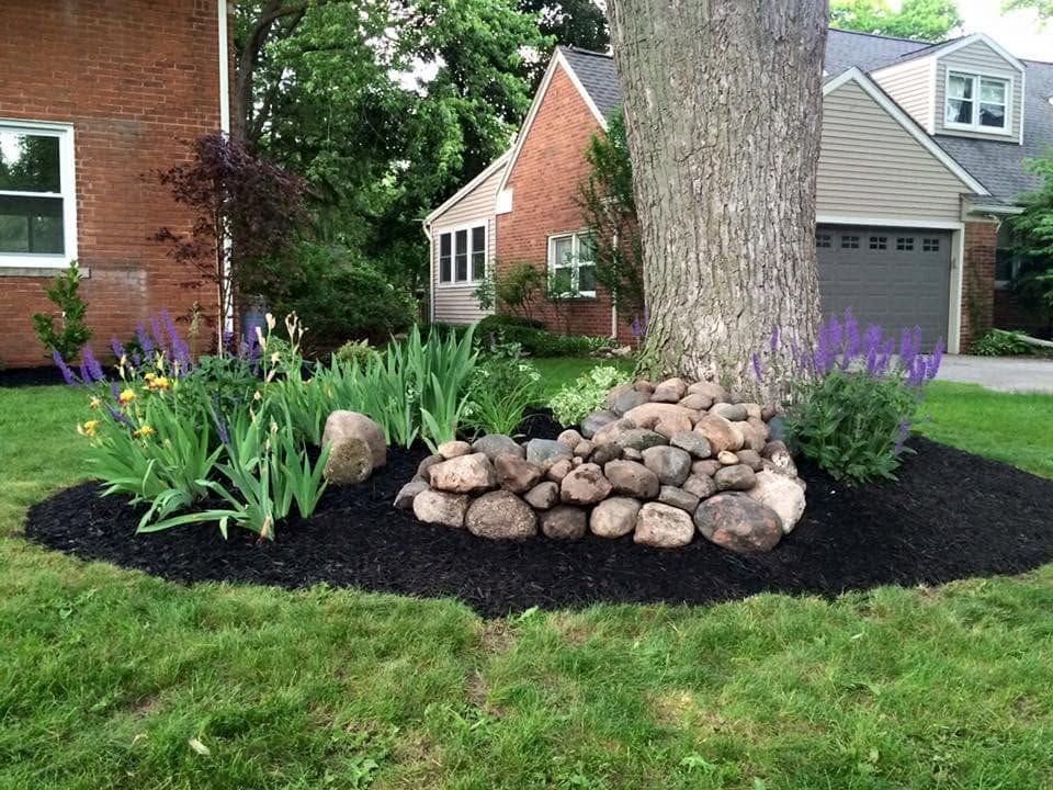 Landscape bed around tree with stacked rocks, flowers, and dark mulch in a suburban yard