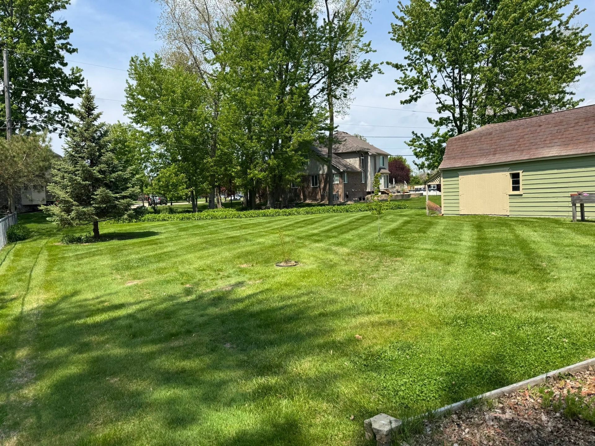 Sunny backyard with green lawn, trees, and a beige house with brown roof.