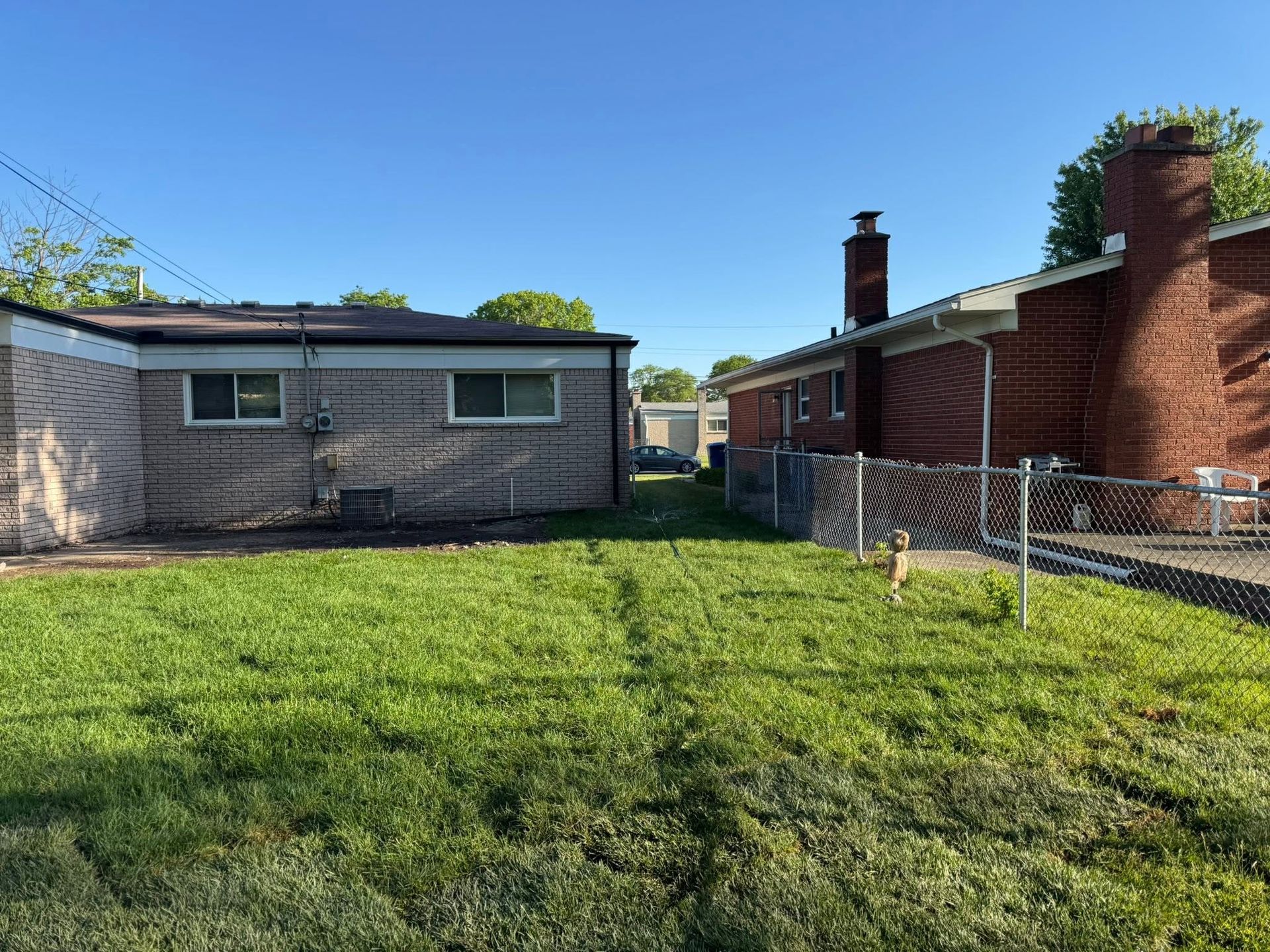 Backyard with green lawn between a gray house and a red brick house, fenced walkway, and clear blue sky