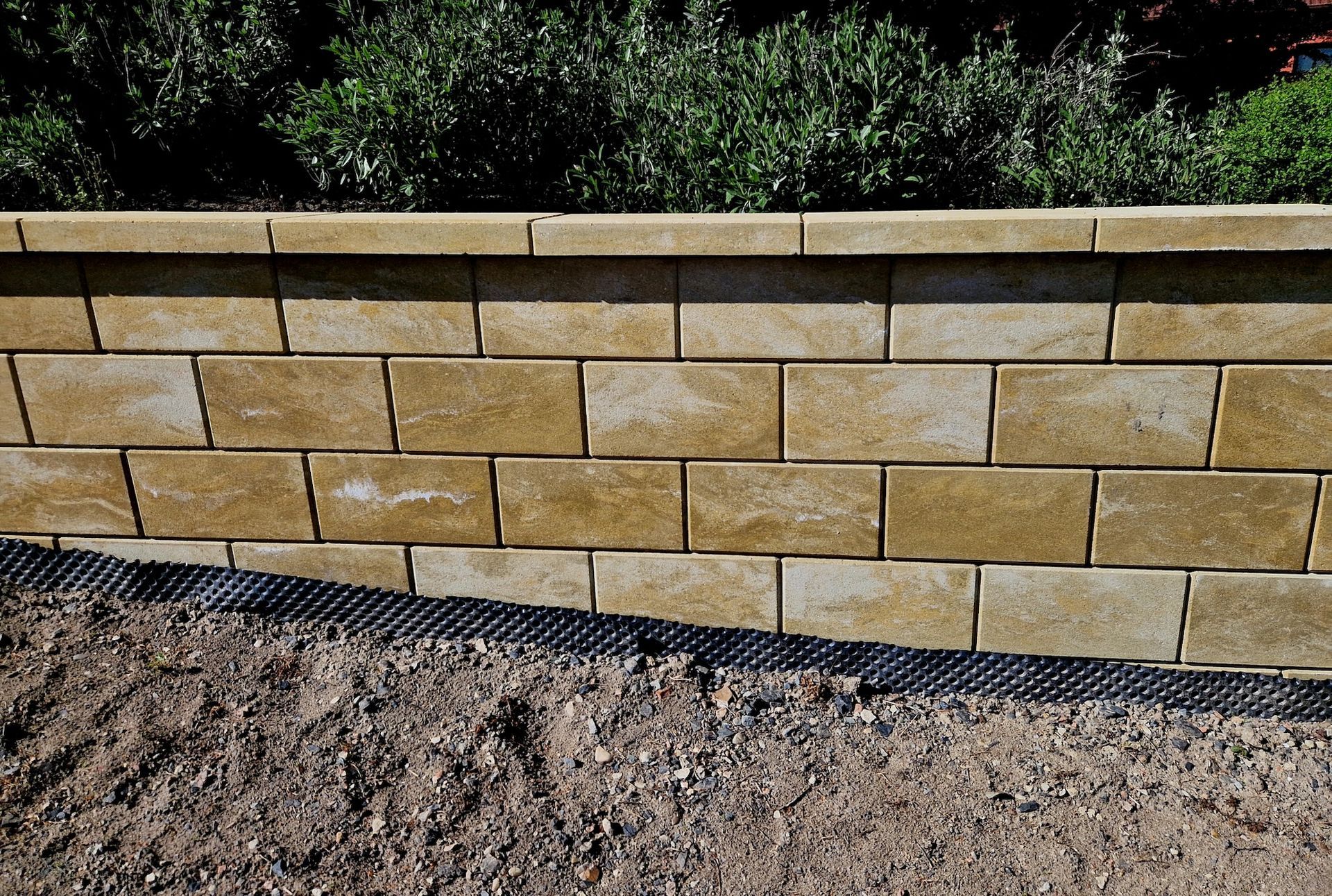 Tan stone retaining wall beside gravel and a black drainage strip in a landscaped area