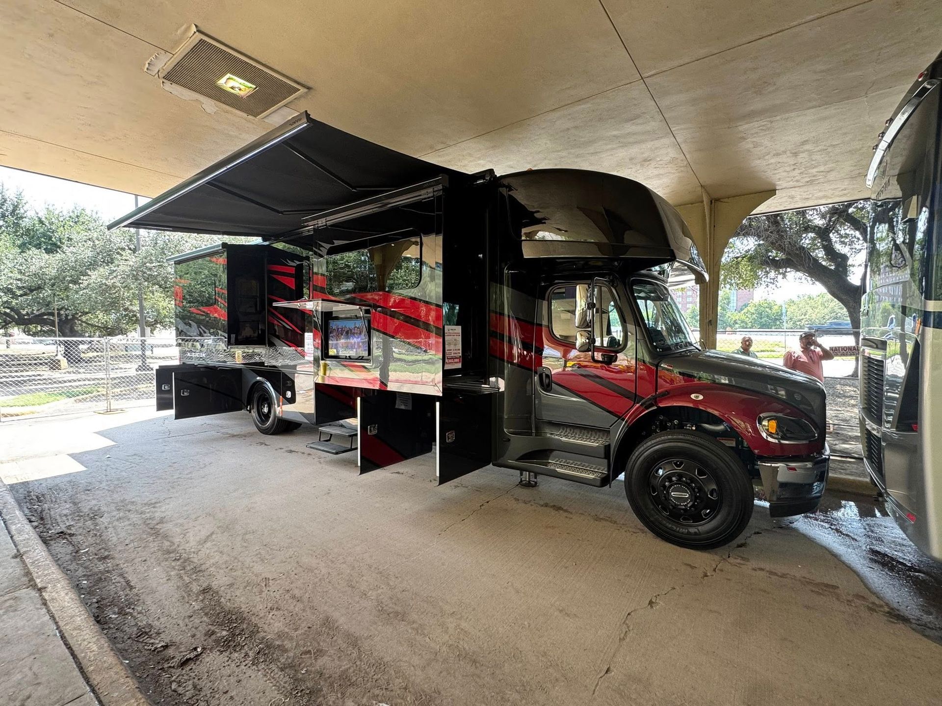 A custom black and red RV parked under a covered area, with open doors and an awning extended.
