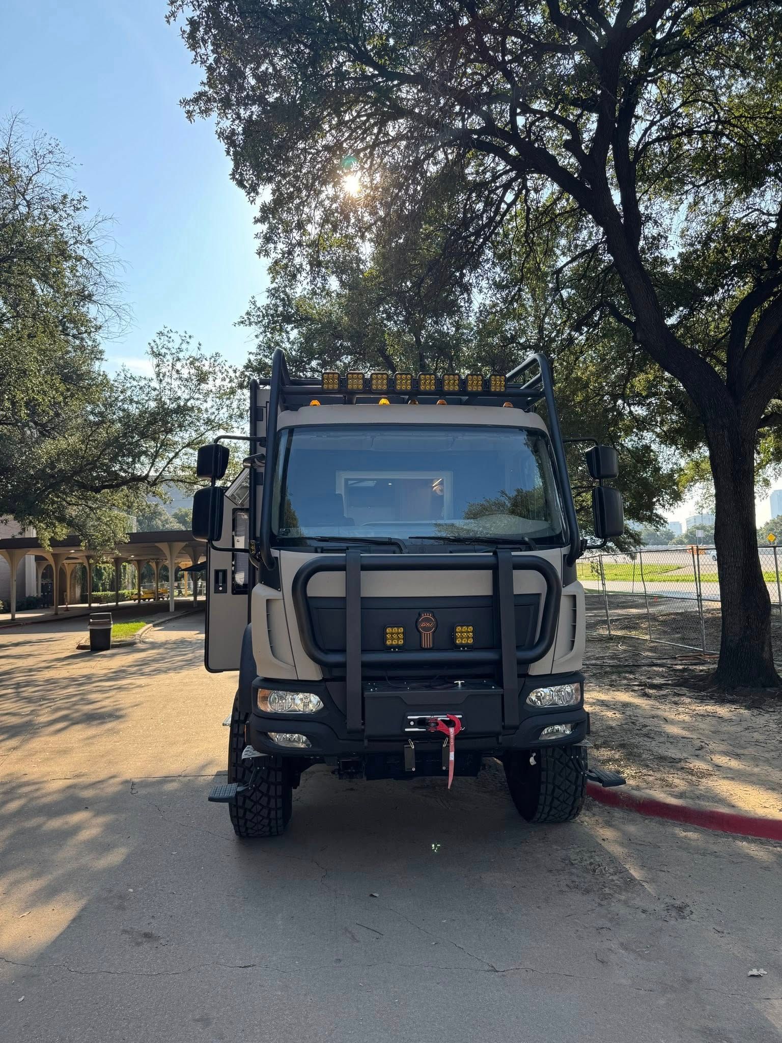 Tan and black custom off-road truck parked on asphalt road, trees in background, sunny day.