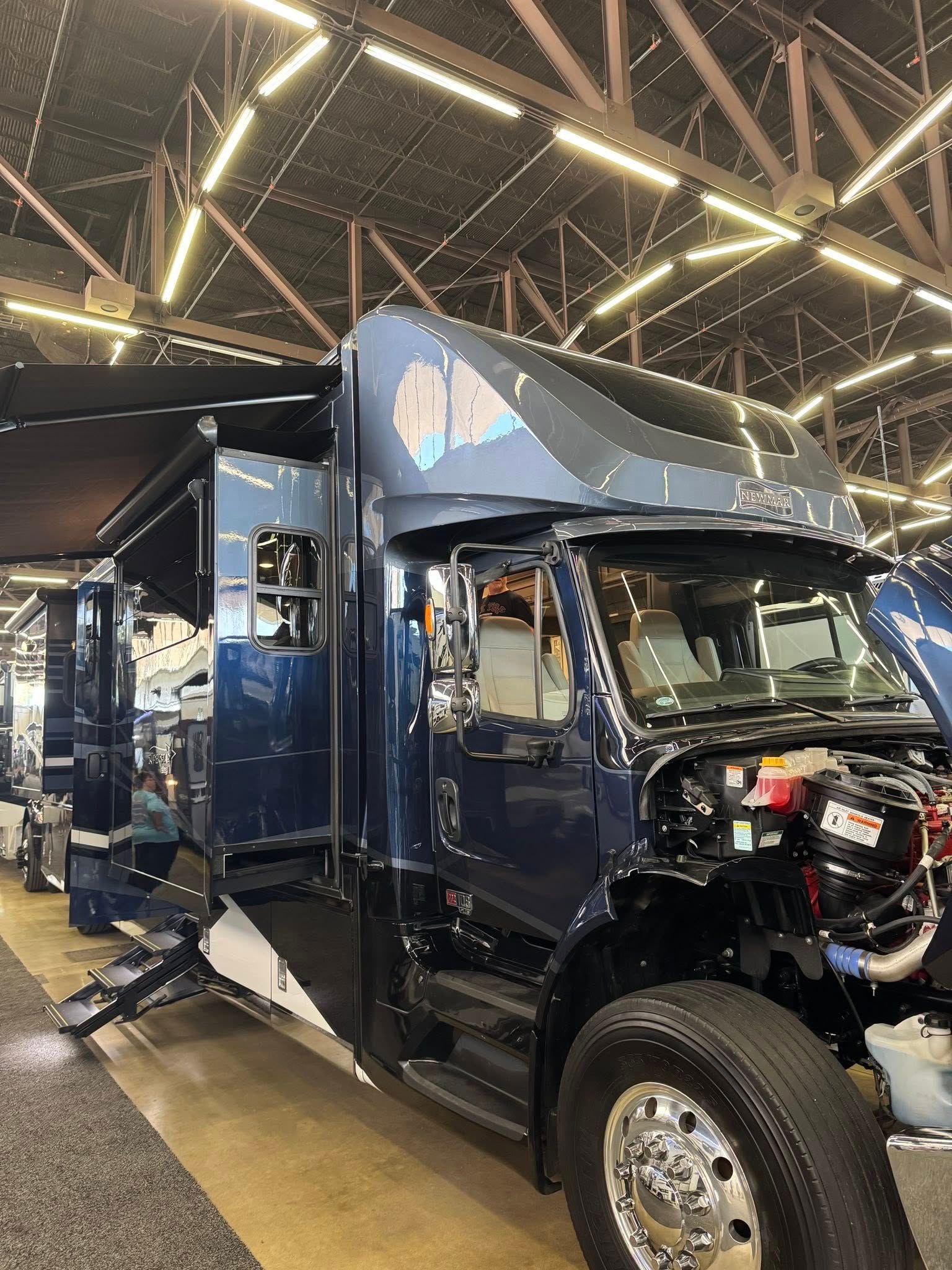 Dark blue RV on display at a showroom, with side stairs extended, engine partially visible.