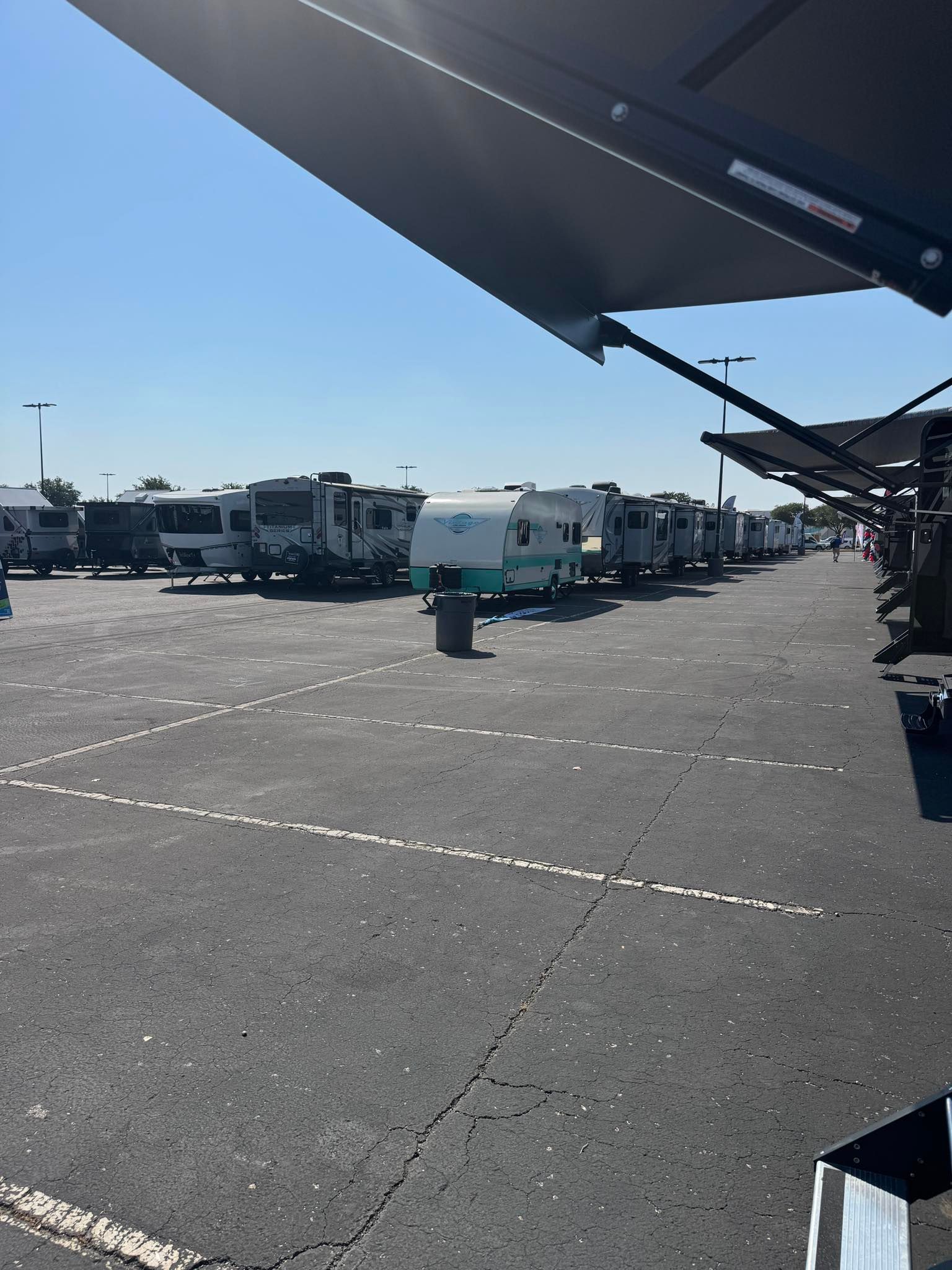 RV campground on a sunny day. Various RVs parked in rows. Awnings provide shade. Asphalt parking area.