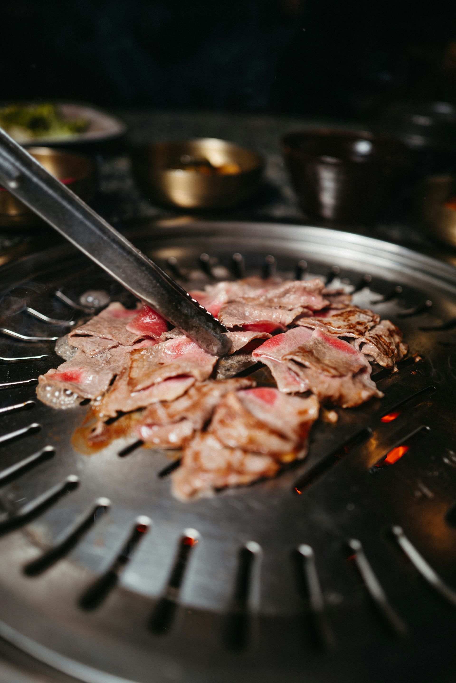 Gros plan sur un barbecue coréen : des pinces retournent de fines tranches de viande sur un gril chaud.