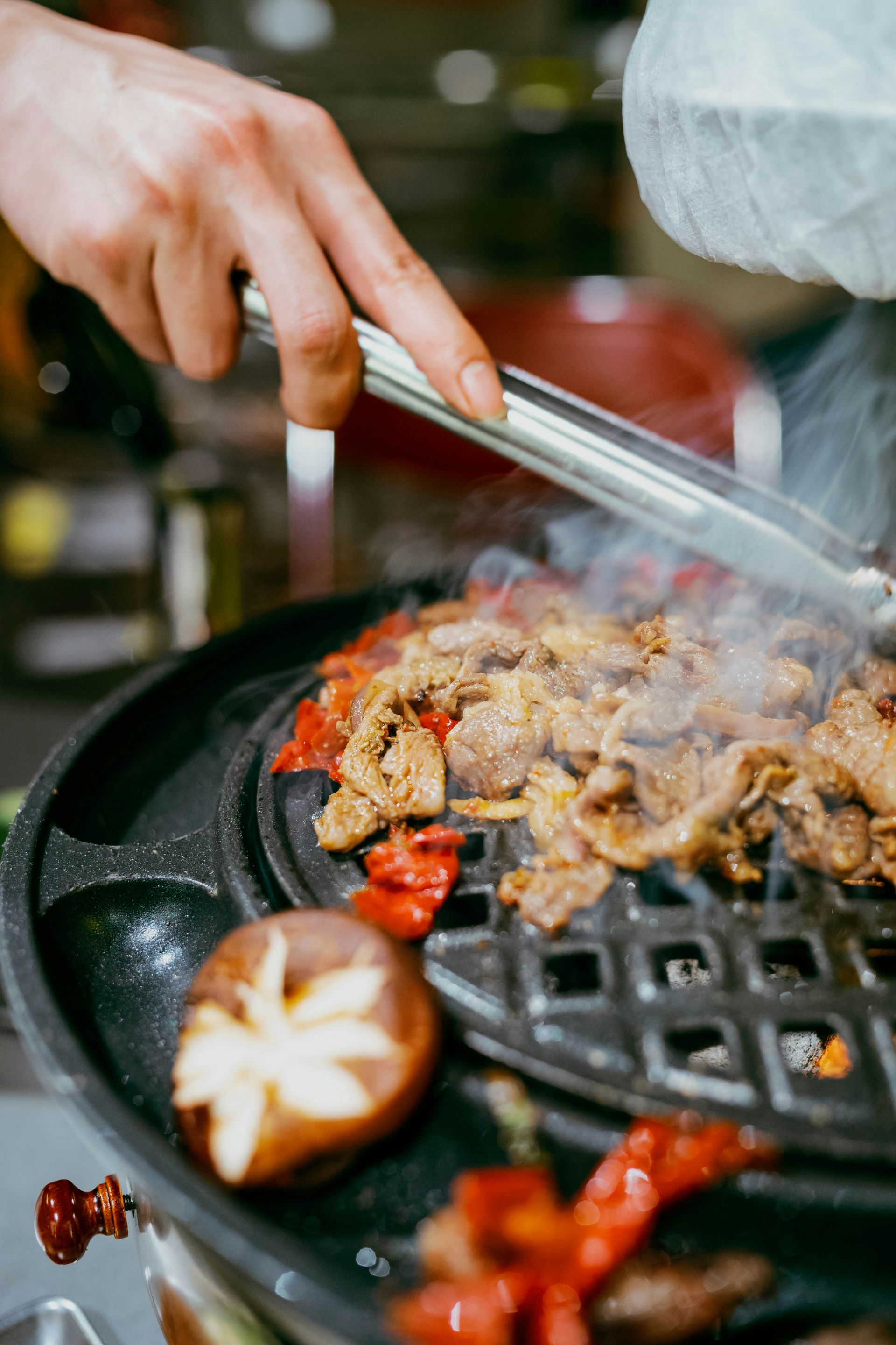 Main utilisant des pinces pour cuire de la viande et des légumes sur un gril, la vapeur s'échappant.