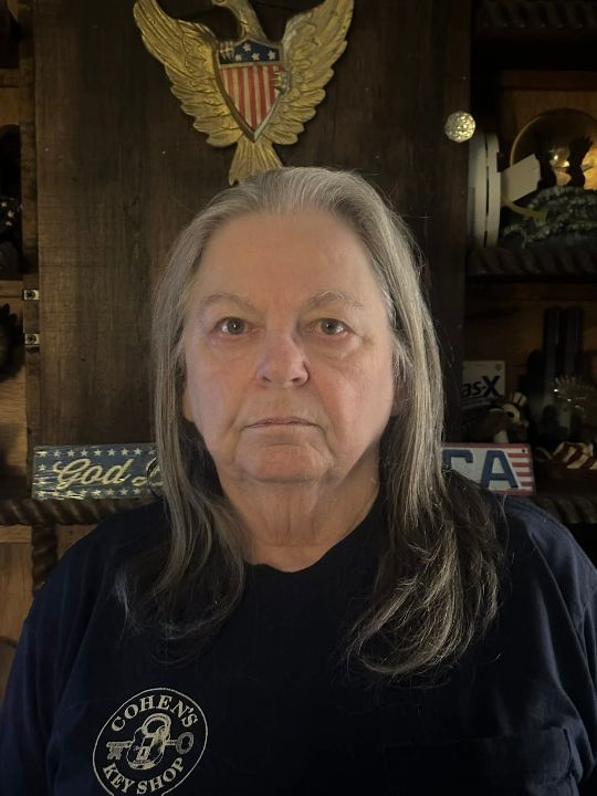 Woman with long gray hair, dark shirt, standing in front of wooden wall with eagle emblem and patriotic decor.