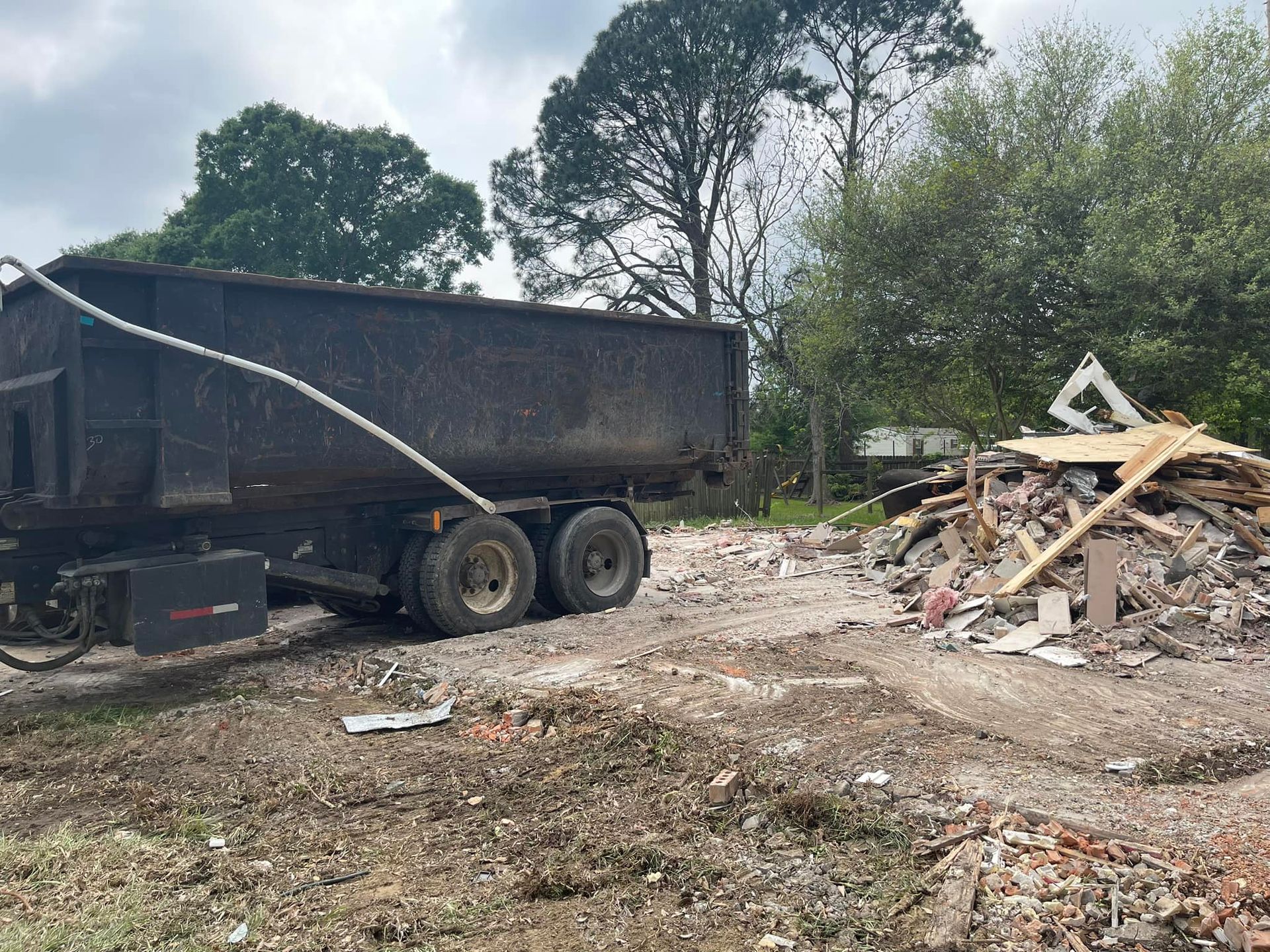 A dump truck is driving down a dirt road next to a pile of wood.