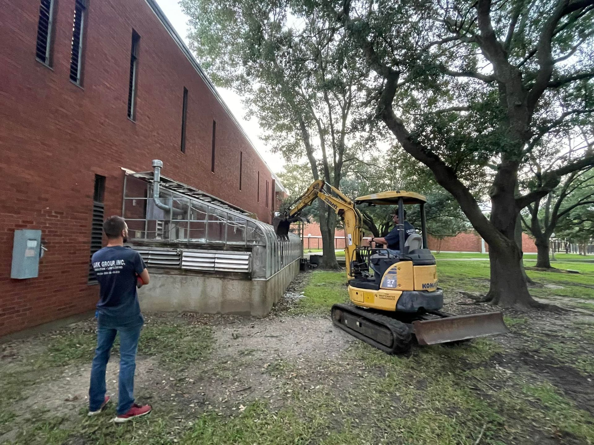 A man is standing next to a yellow excavator in front of a brick building.