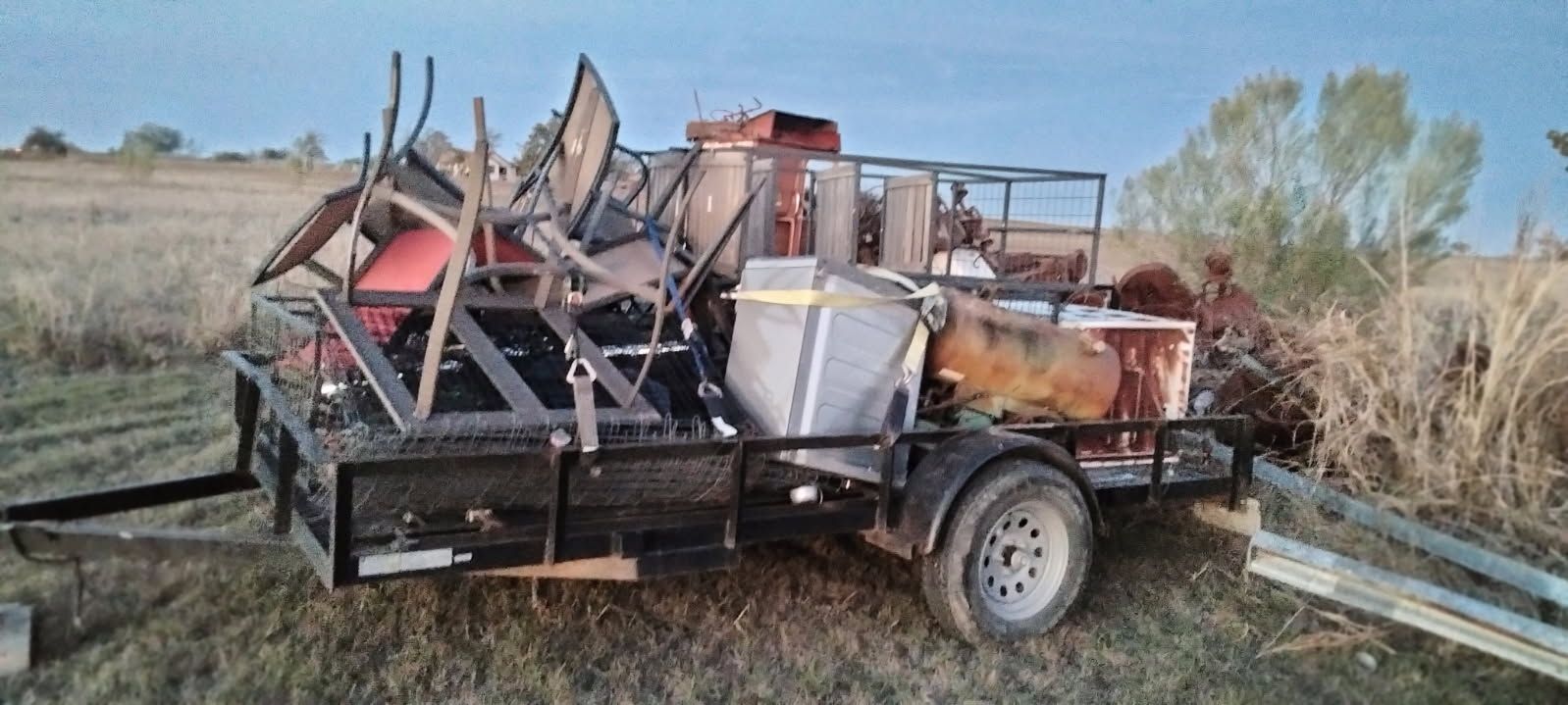 Trailer loaded with metal farm equipment and debris in a grassy field under a clear sky