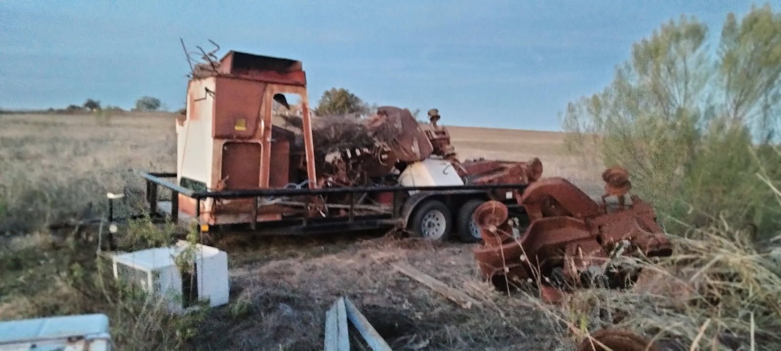Rusty old farm machinery loaded on a trailer in a dry field, with scattered equipment in the foreground.