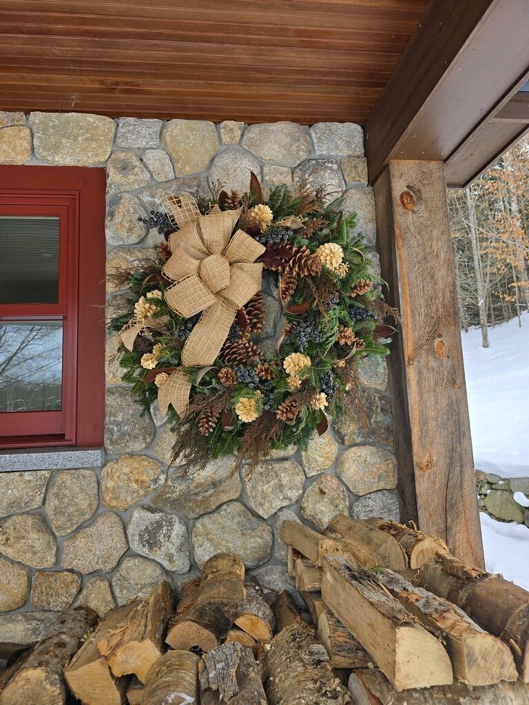 Stone cabin wall with a large burlap holiday wreath beside a red window and stacked firewood in snow