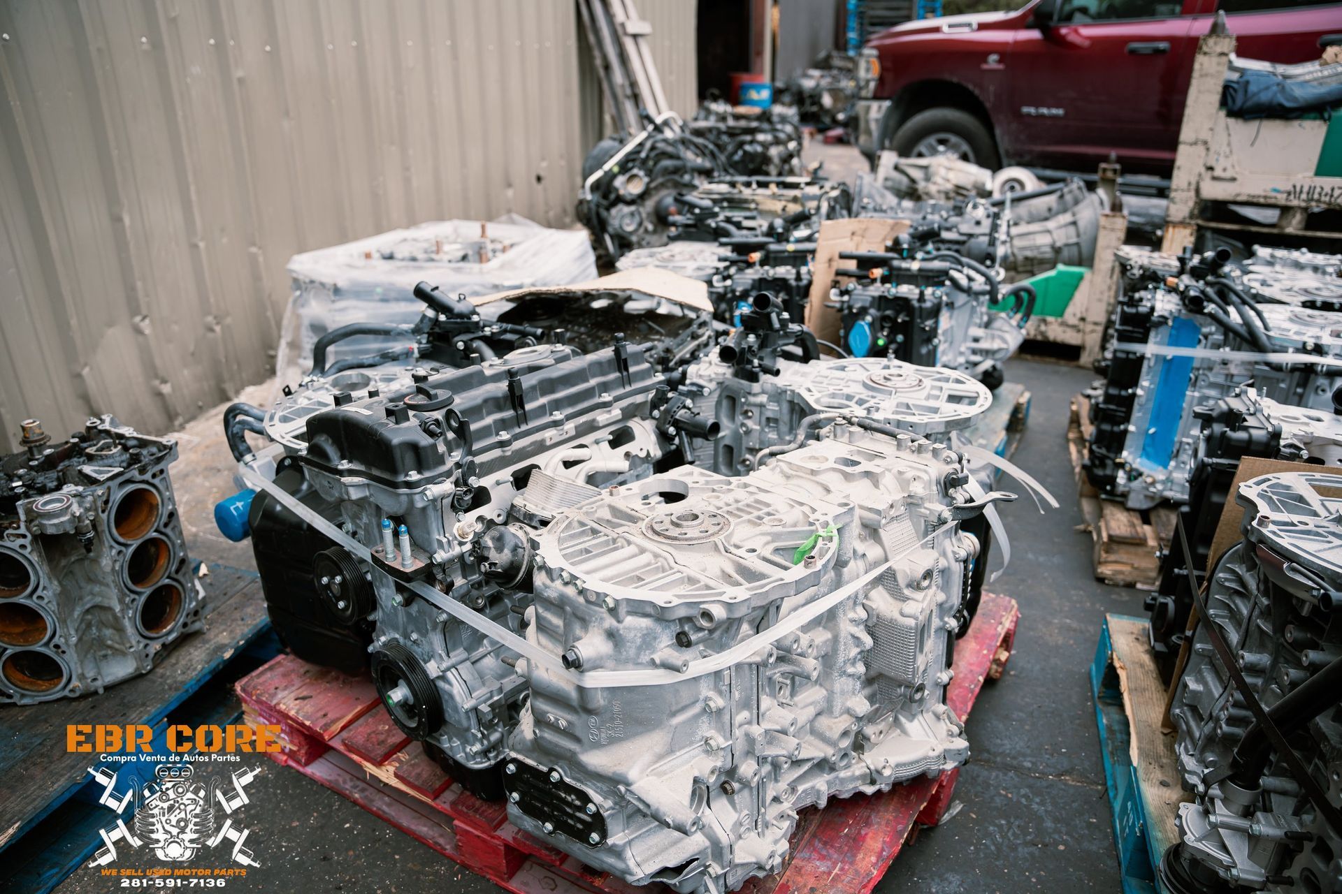 Pile of car engines, some on pallets, outdoors near a building and a red truck.