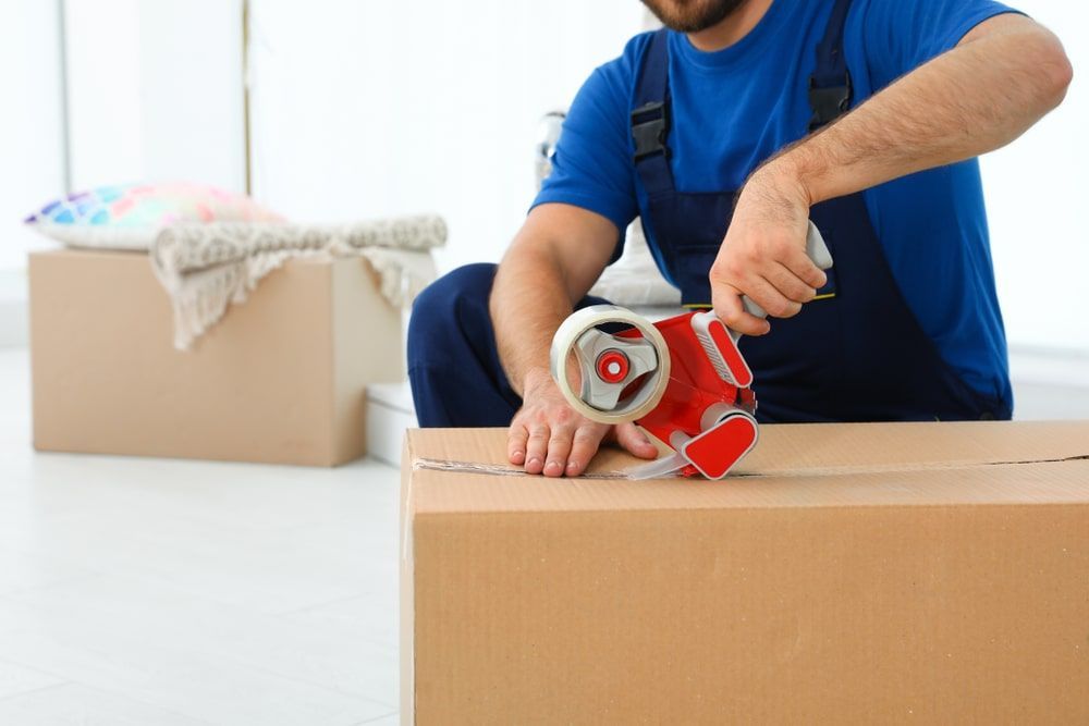 A Man Is Sitting On A Box Using A Tape Dispenser — Greater Newcastle Removals In Rathmines, NSW