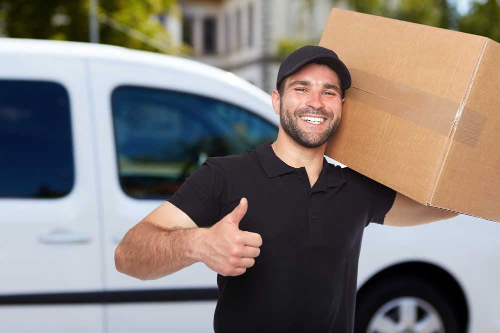 A Delivery Man Is Holding A Cardboard Box And Giving A Thumbs Up — Greater Newcastle Removals In Swansea, NSW