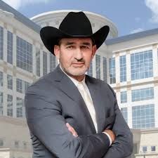 A man in a suit and cowboy hat is standing in front of a building with his arms crossed.