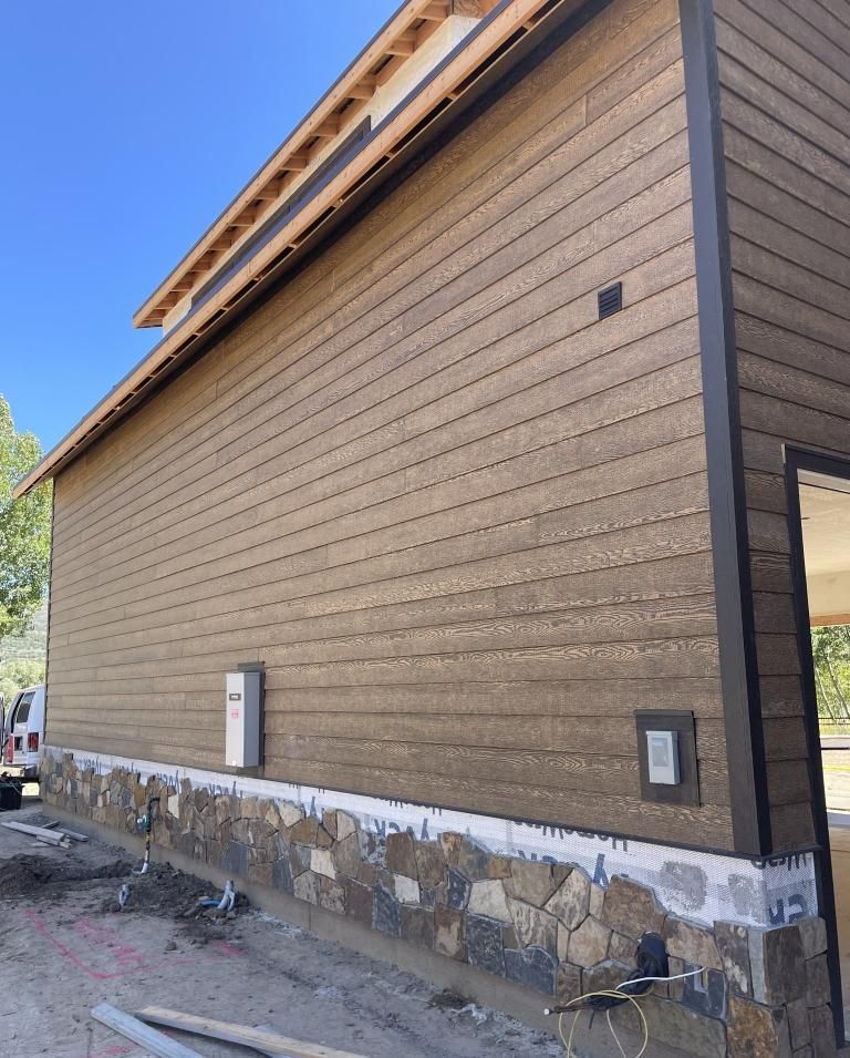 Brown building exterior with horizontal siding, stone base, and dark trim against a blue sky.