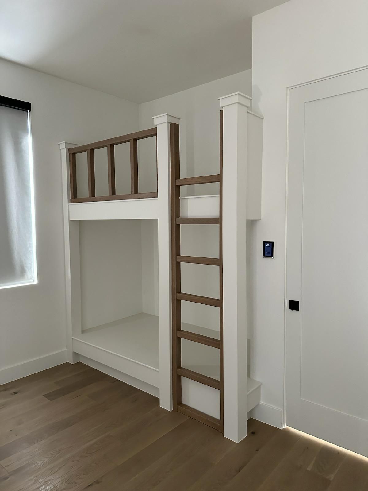 Bunk bed in a white room with a wooden ladder and trim. Light hardwood floor.