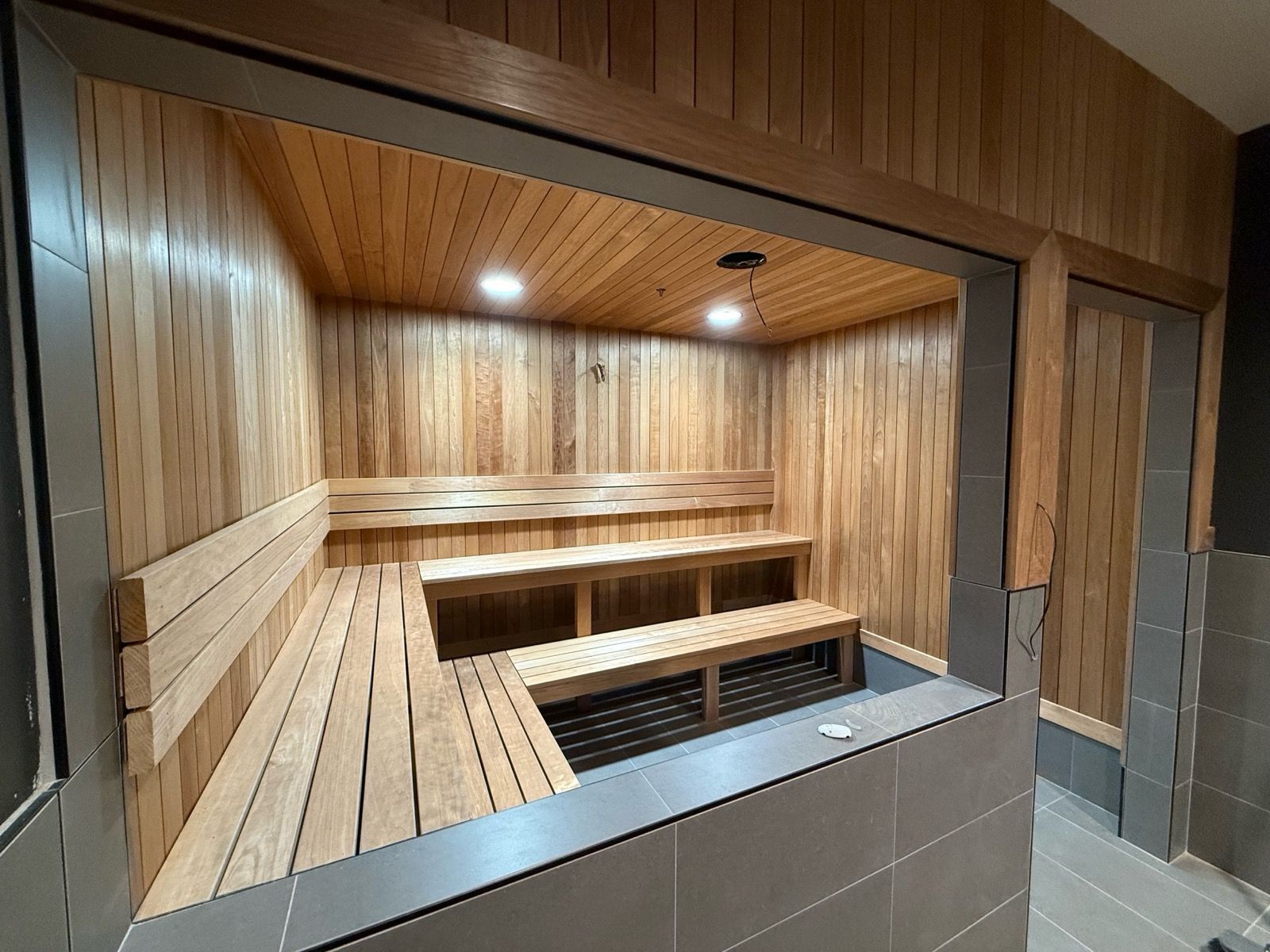 A cedar wood sauna with multiple benches and recessed lighting. Tile exterior framing the opening.
