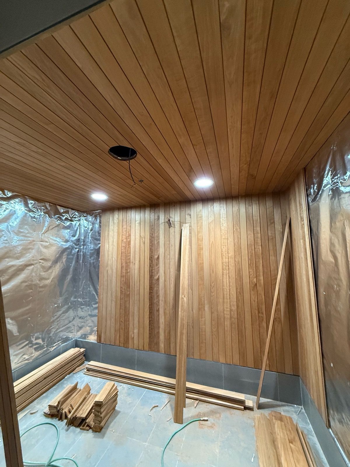 Wooden planks installed on walls and ceiling of a room, with recessed lighting and protective covering.