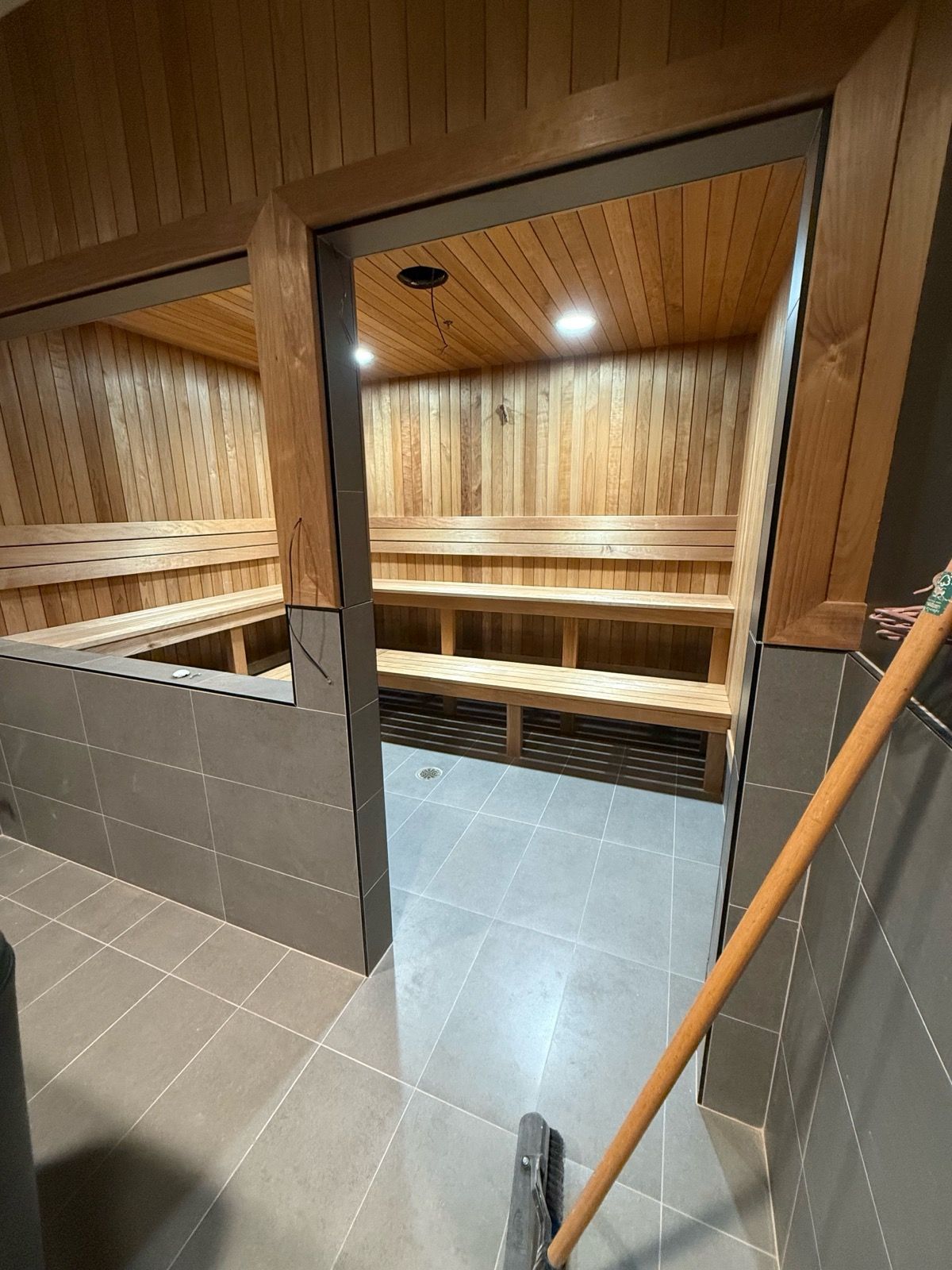 Two cedar-lined saunas with benches, accessed through a doorway and separated by tiled walls. A broom leans against the wall.