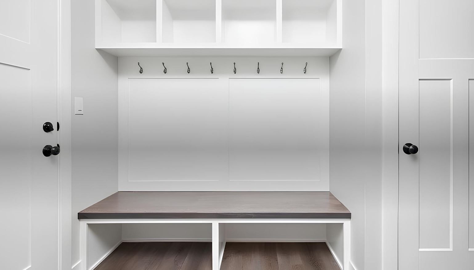 White mudroom bench with hooks and overhead cubbies between two doors, gray wall and wood floor