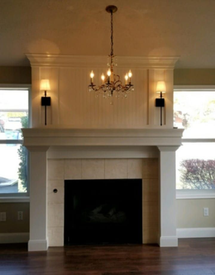 Elegant living room fireplace with chandelier, wall sconces, and large windows on either side.