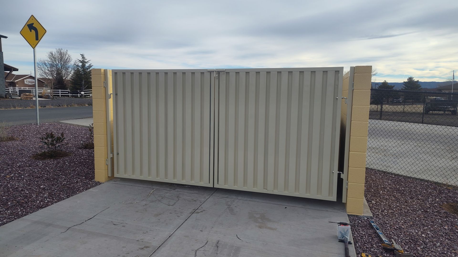 Tan corrugated metal gate, closed, between tan brick pillars. Asphalt driveway, rocky ground, road sign, cloudy sky.