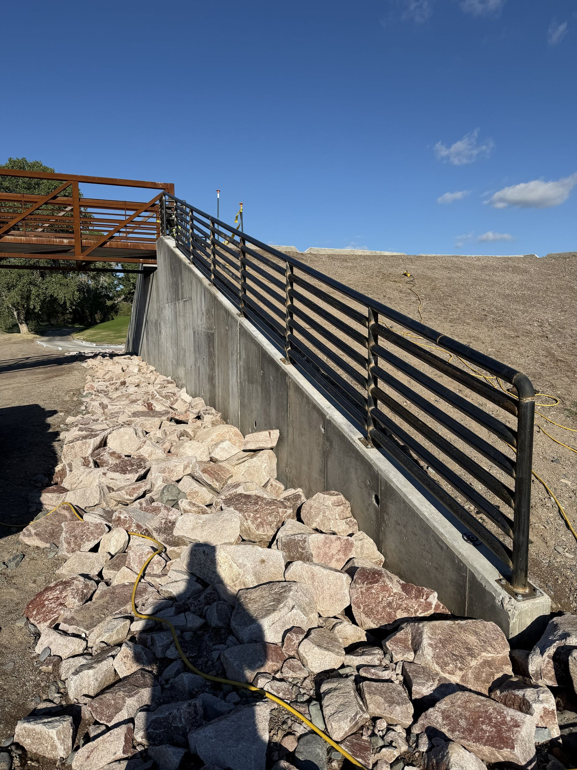 A concrete retaining wall with a black metal handrail runs up a gravel slope toward a wooden bridge under a blue sky.