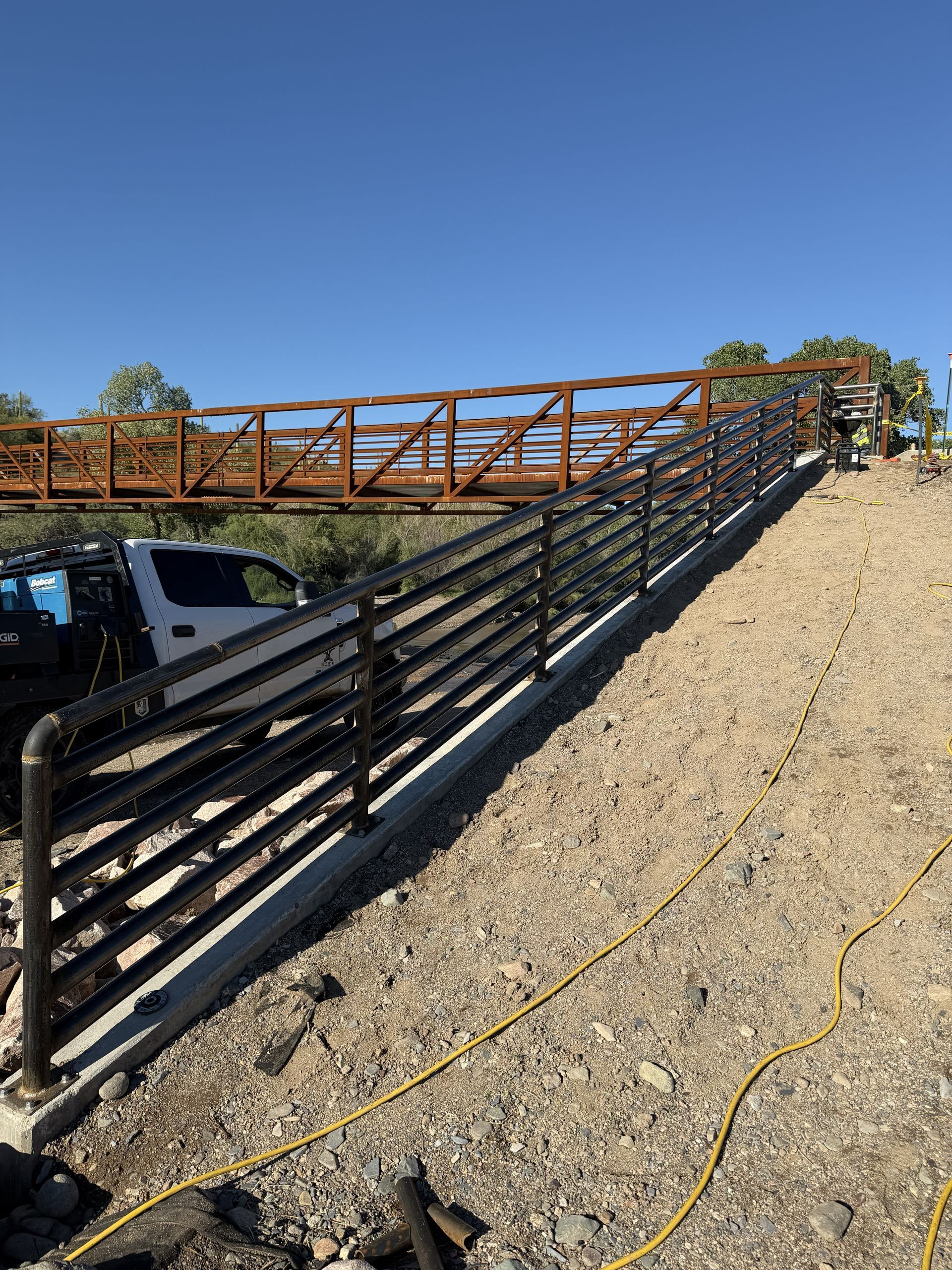 A white utility truck parked beside a metal railed ramp leading to a bridge under construction on a gravel slope.