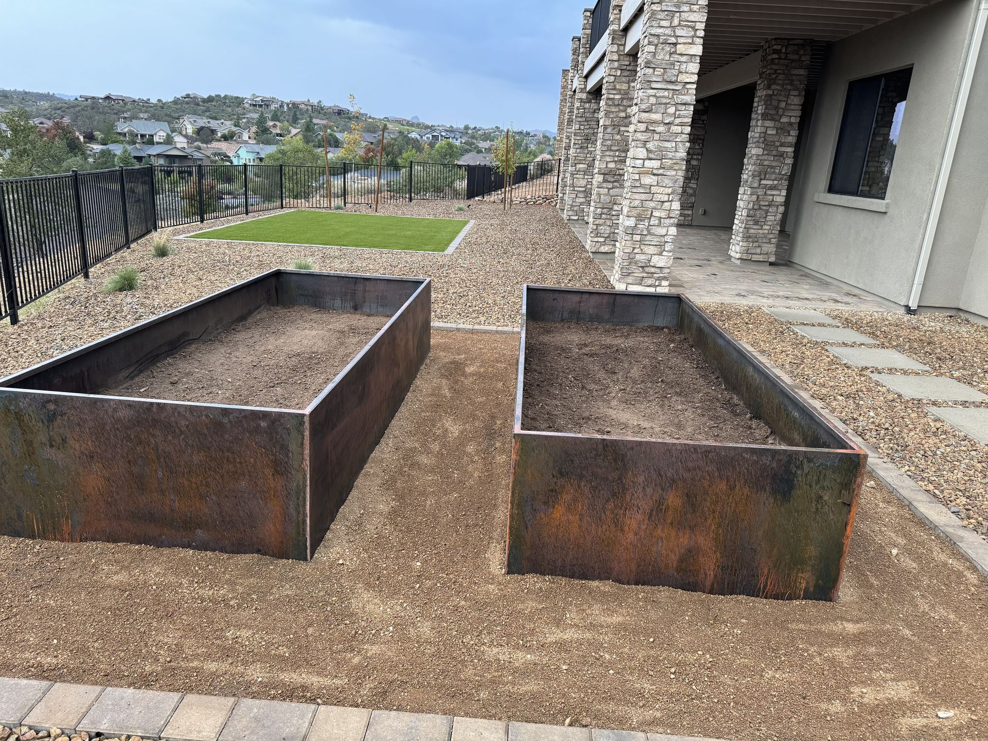 Two rectangular, rusted metal raised garden beds sit on a gravel ground next to a stone-clad building and a lawn.