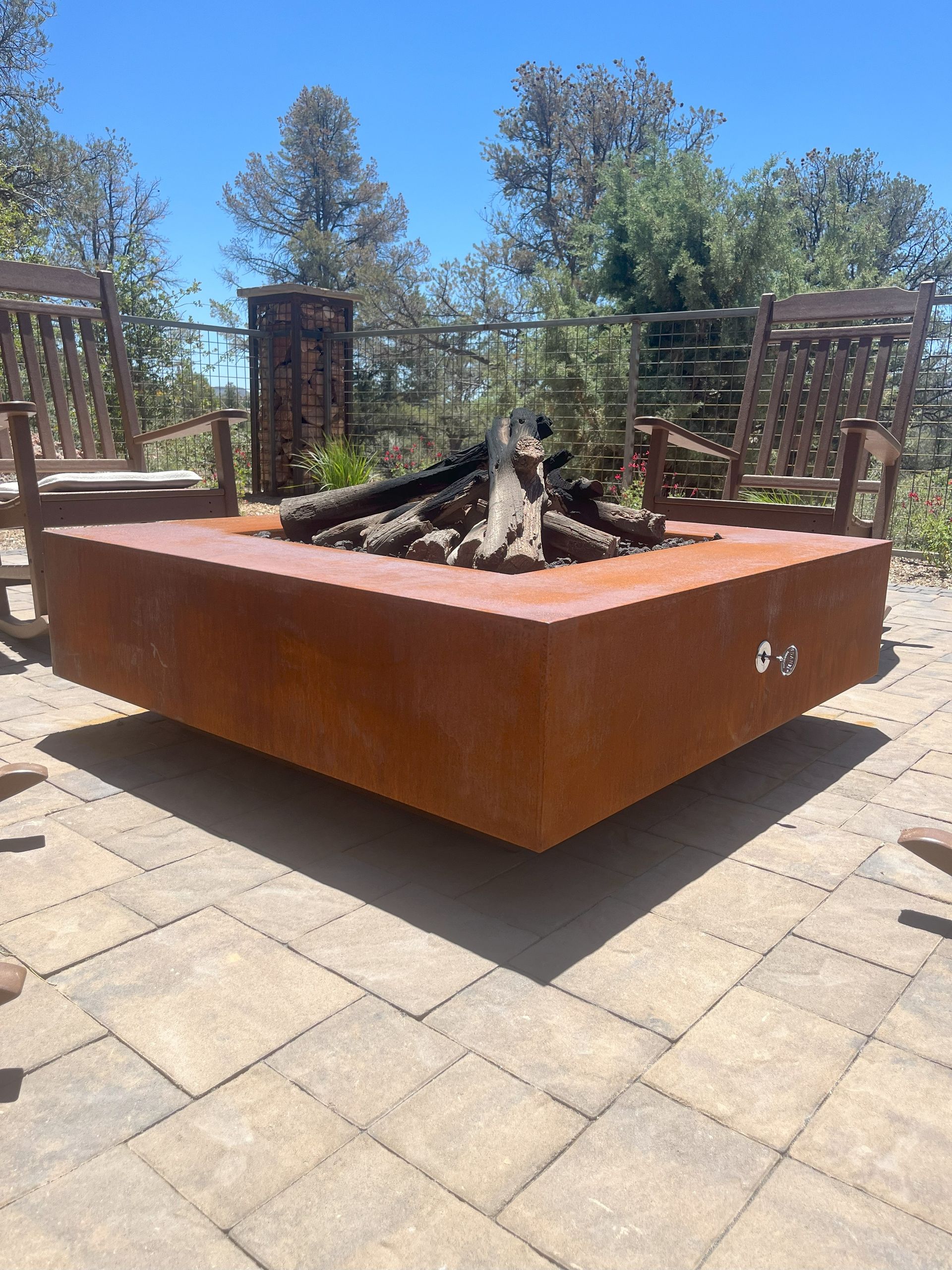 Rust-colored square fire pit with burning logs on a stone patio, wooden chairs in background.