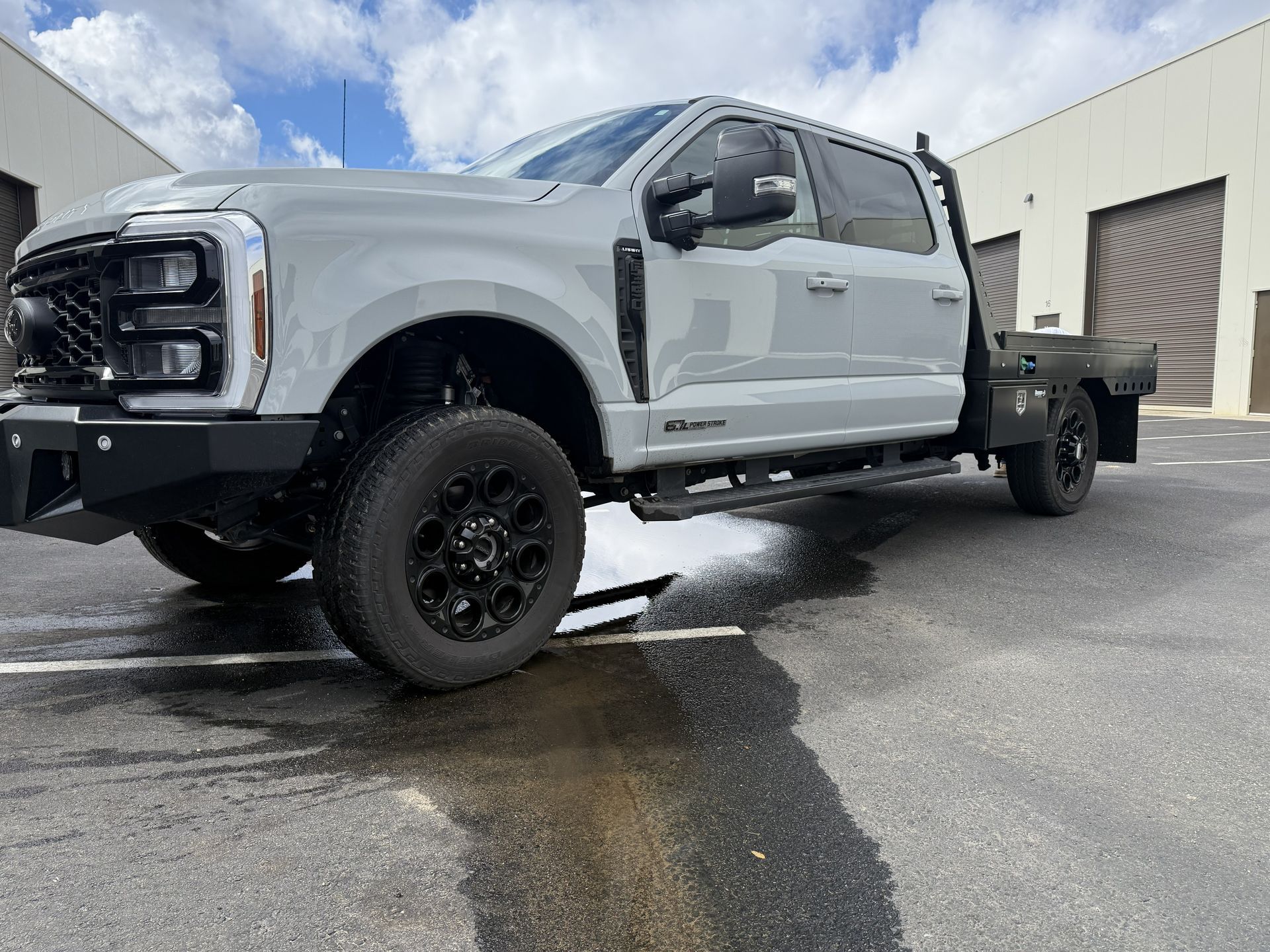 A white Ford F-Series pickup truck with a flatbed body parked on asphalt in front of two industrial buildings.