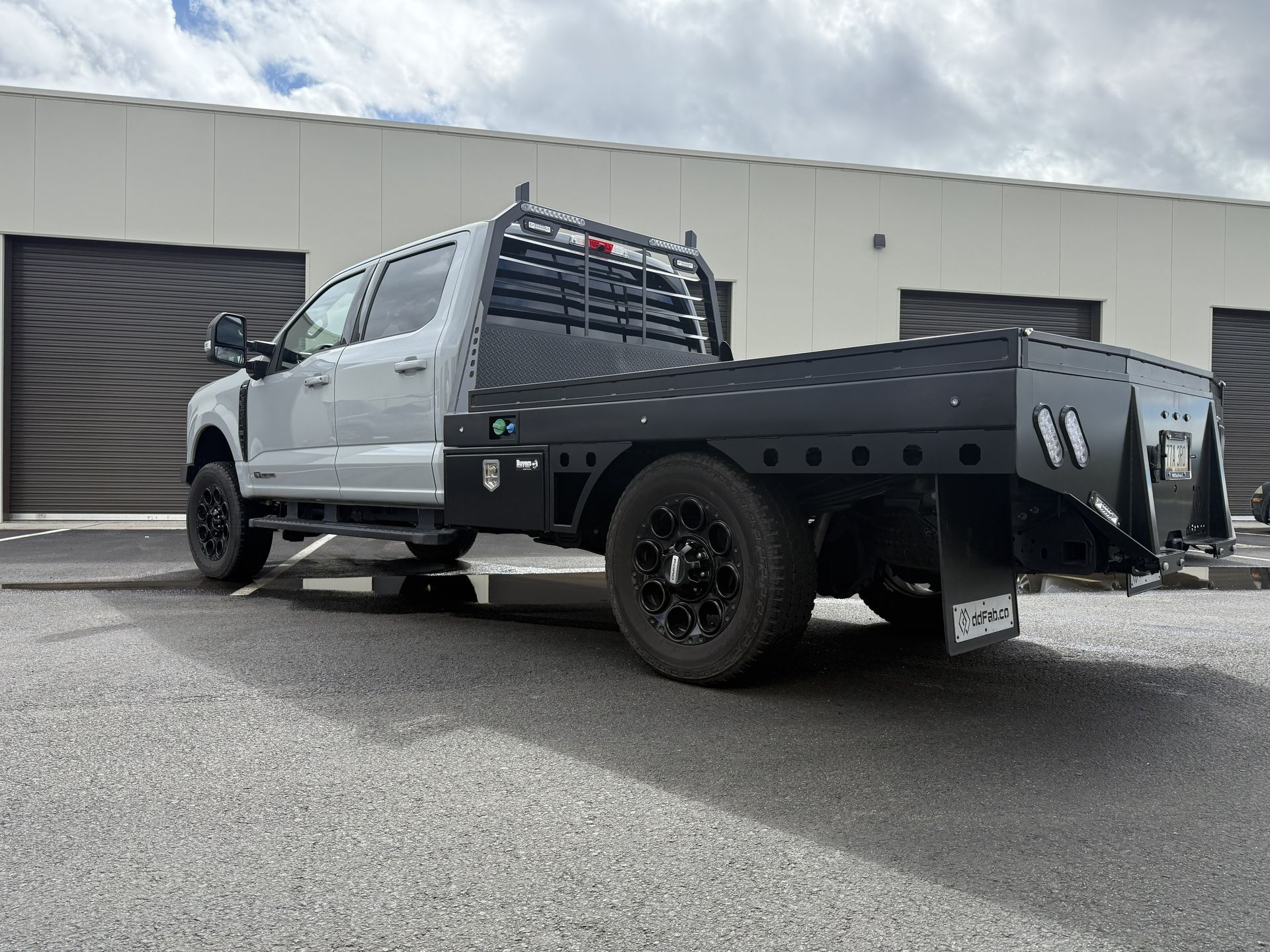 Silver crew cab pickup truck with a black flatbed utility body parked in an outdoor lot against a building.