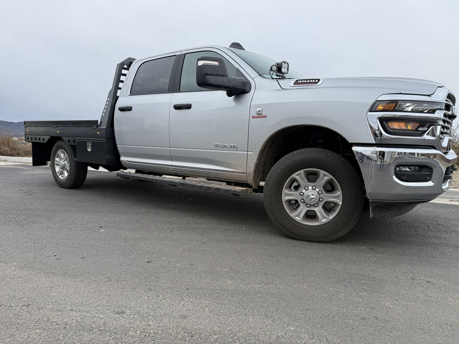 Silver Ram 2500 crew cab truck with a flatbed utility bed parked on a gravel surface under a cloudy sky.