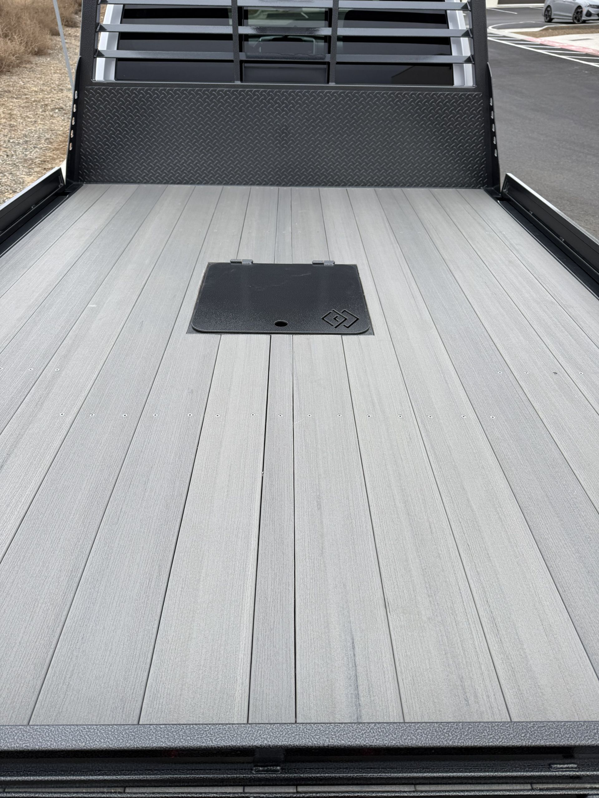 A view looking down at a flatbed truck bed featuring light gray wood-look planks, a black metal bulkhead, and a center hatch.
