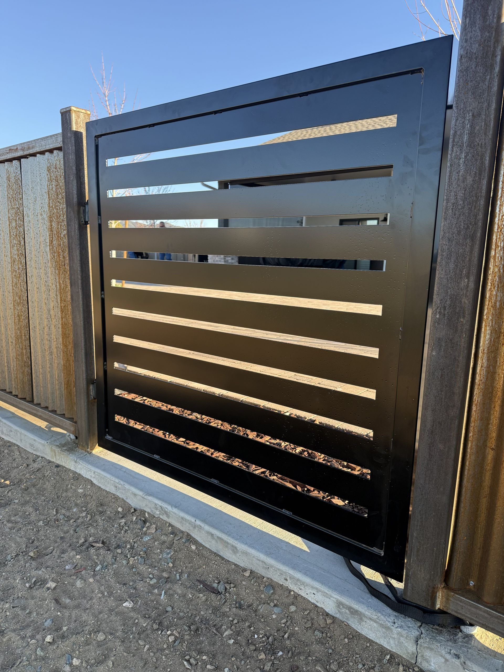 A black, horizontal-slatted metal pedestrian gate installed between wooden fence posts on a gravel ground surface.