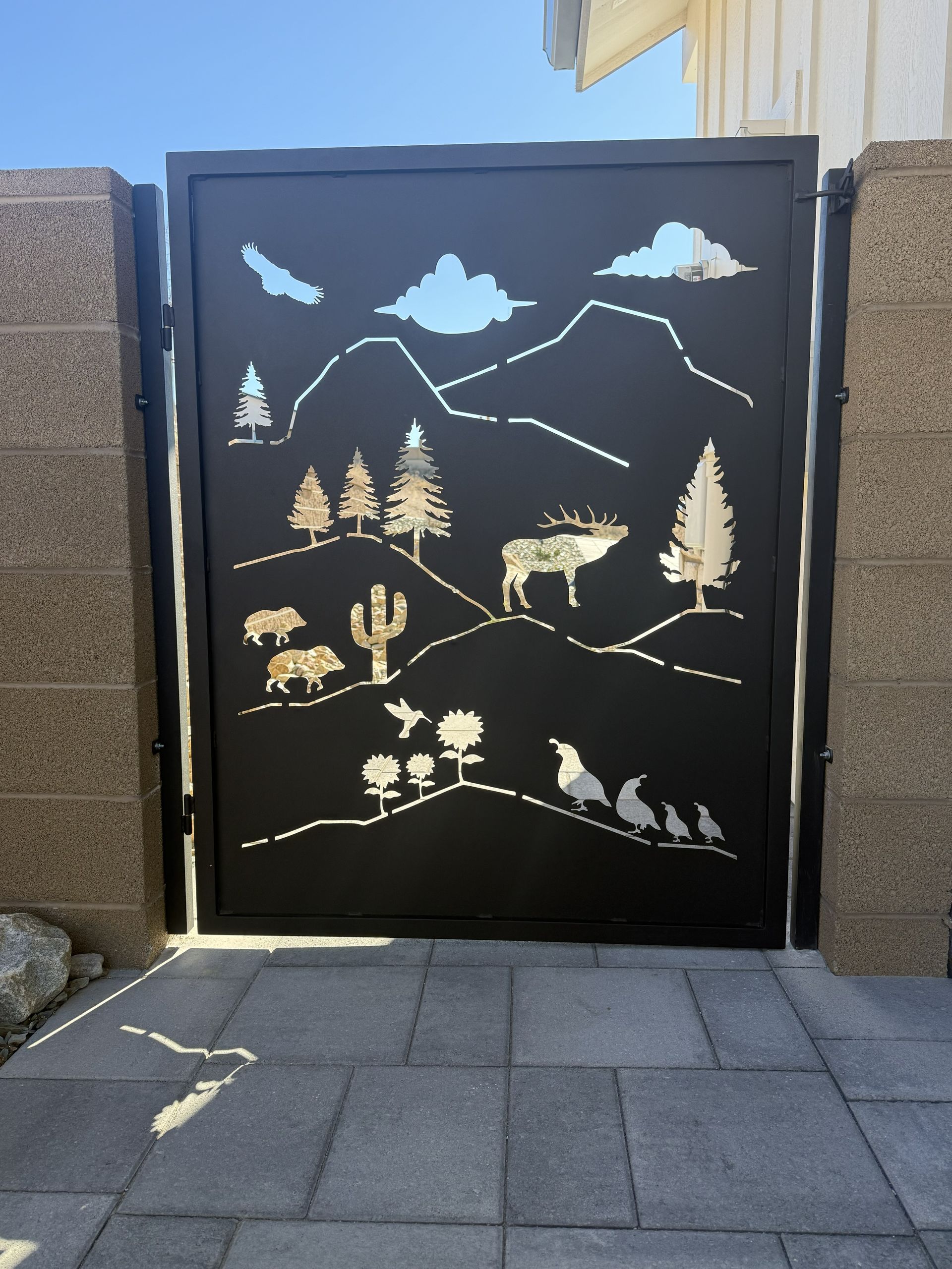 Black metal gate featuring laser-cut silhouettes of desert mountains, pine trees, an elk, a cactus, and small animals.